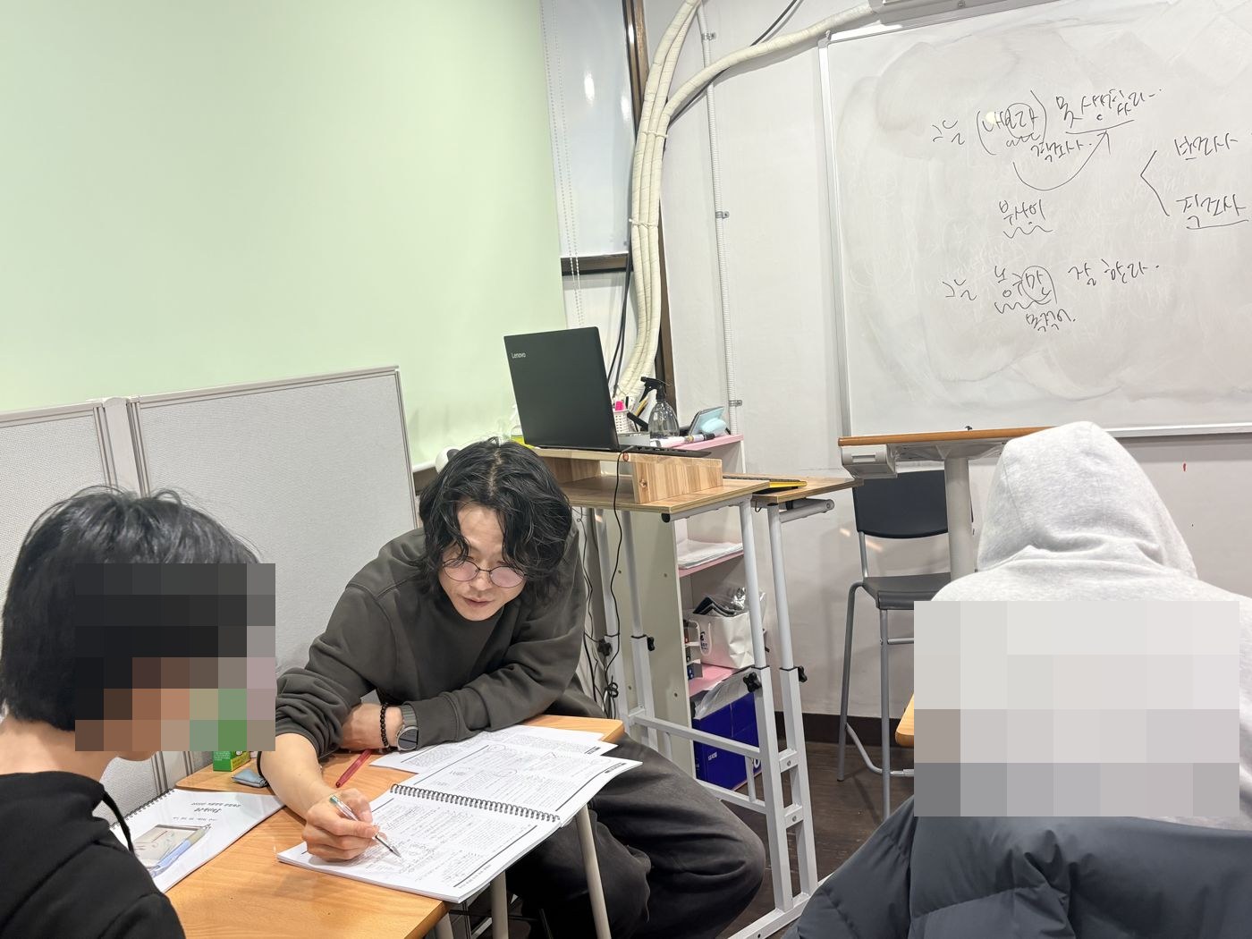 화이트보드에 문법 도식을 써 둔 교실에서 원장이 학생과 마주 앉아 교재에 첨삭하는 모습